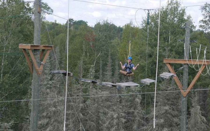 an outward bound student sits cross-legged while being suspended mid-air during a ropes course